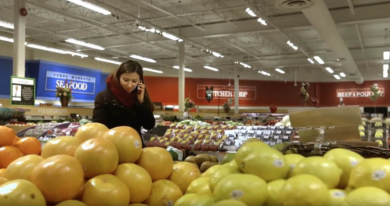 A video thumbnail of a screen capture from the 'IFCO: let's eat' video. The capture features a women shopping in the fruit section of a supermarket. In the foreground of the capture is a bunch of fruit.