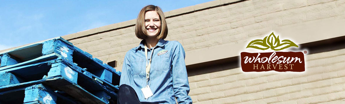 A banner image of a photograph of a happy caucasian women, Wholesum Harvest employee, sitting on a stack of CHEP blue pallets. Beside the women is the Wholesum Harvest logo.