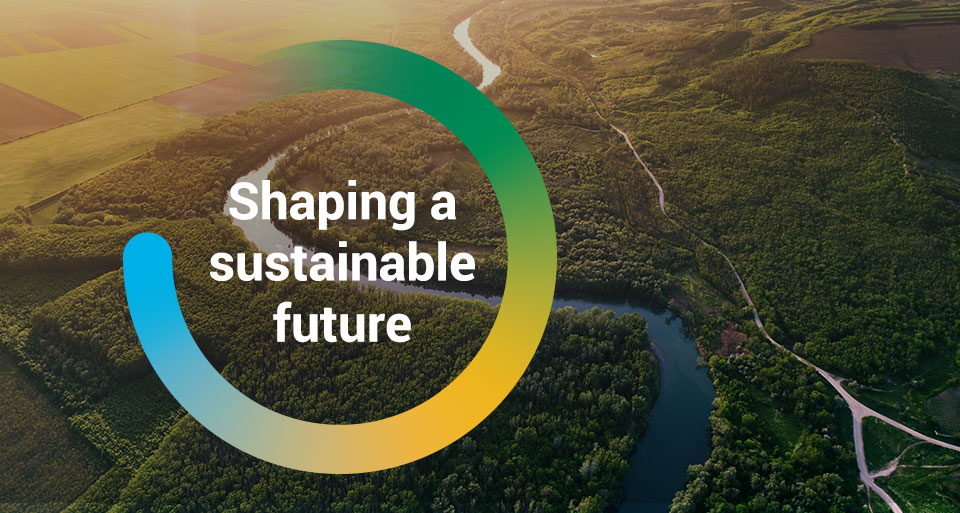 The wordAn aerial photograph of a river with the words Shaping a Sustainable Future inside a colourful circles, 'Shaping our Future' inside a circle graphic overlayed on top of an image of a moving pallet on a conveyor belt